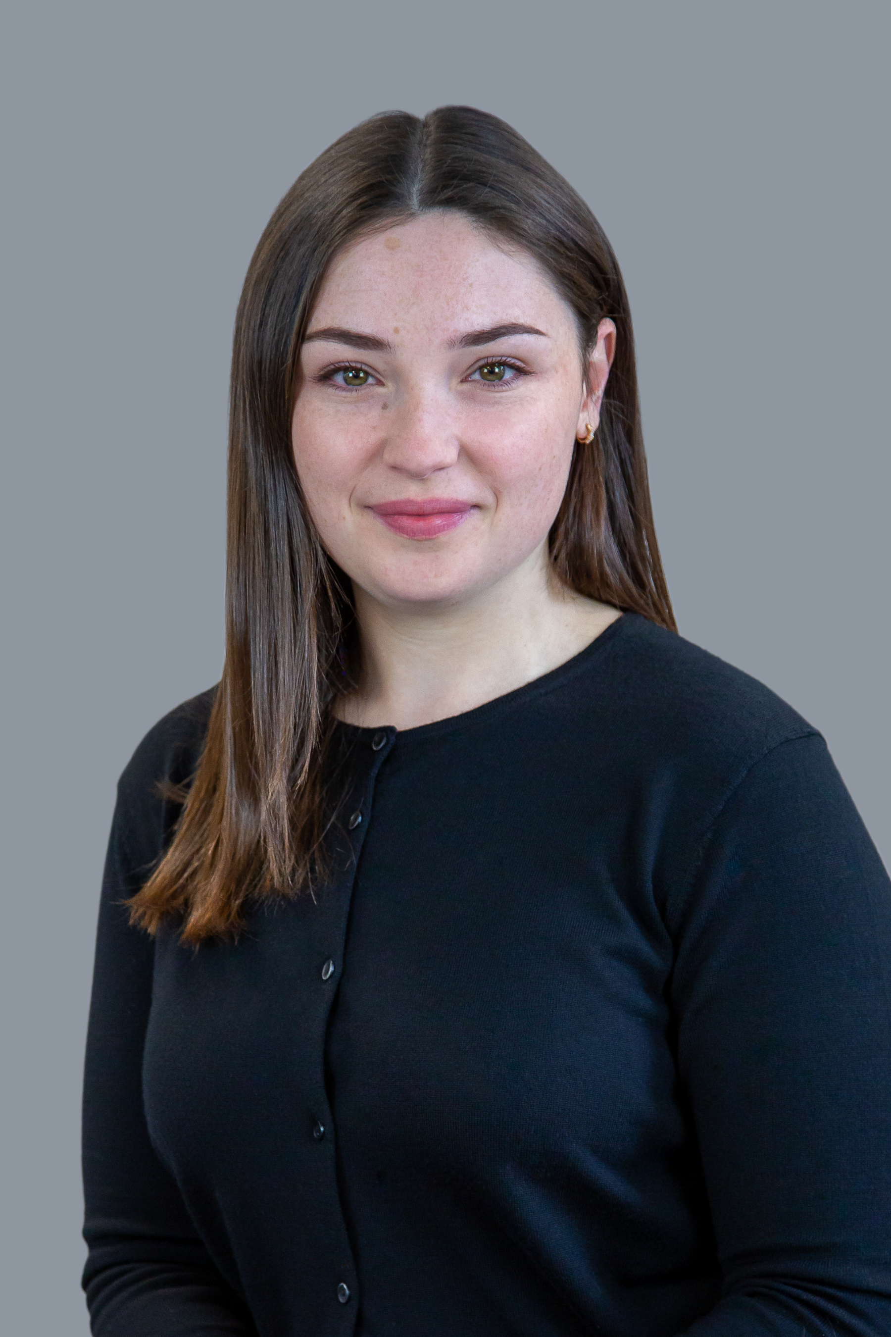 A person wearing a black buttoned top stands in front of a plain grey background and looks at the camera with a slight smile.