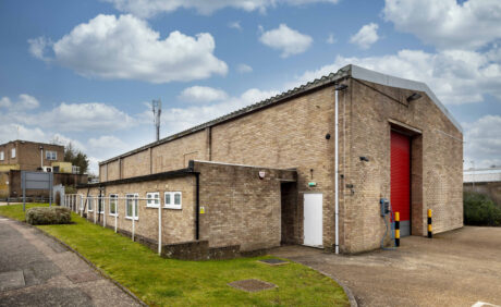 A large industrial brick building with a sloped metal roof, red roller shutter door, small windows, and surrounding paved and grassy area under a partly cloudy sky.