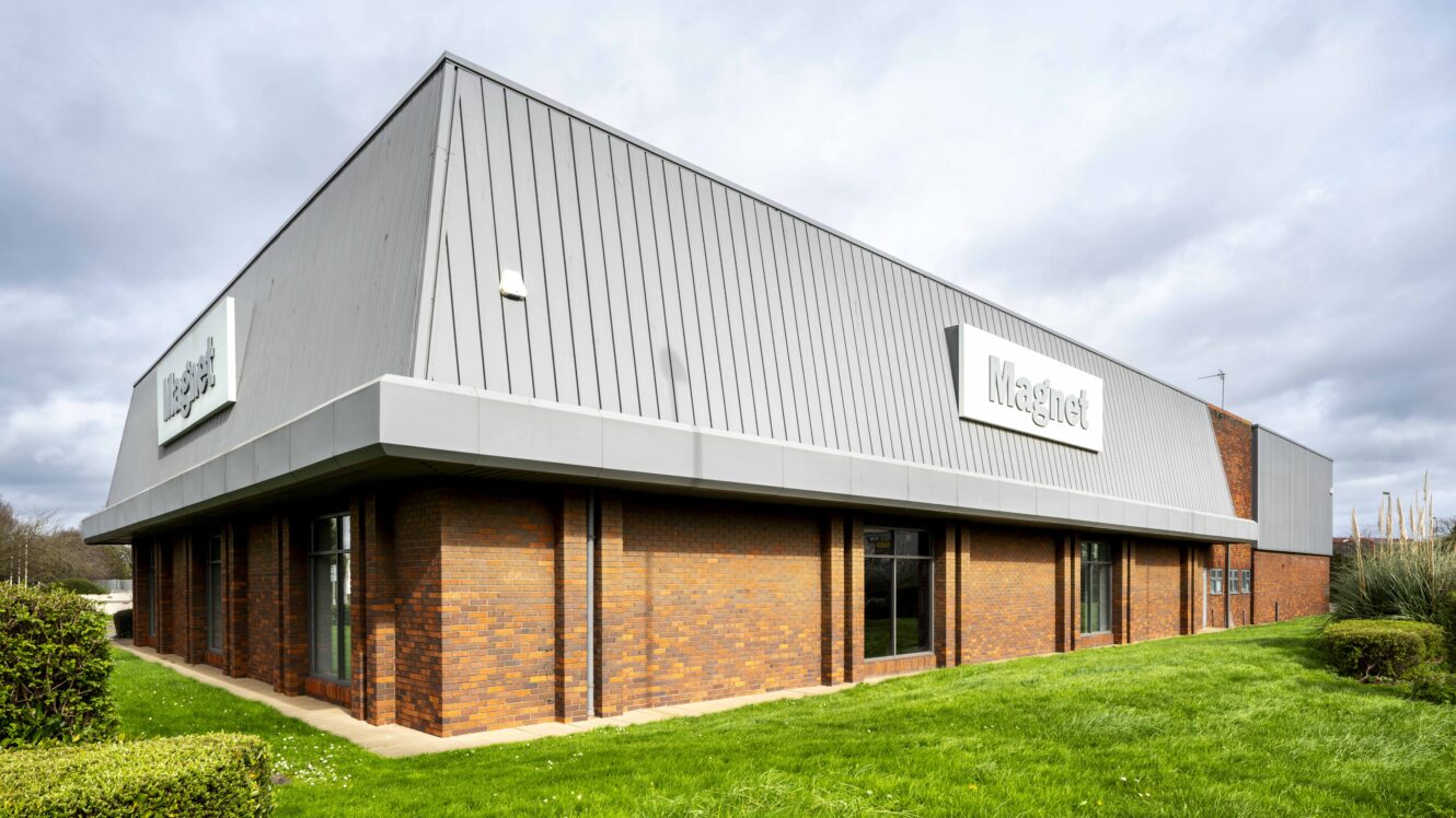 A modern commercial building with brick walls and large windows, featuring “Magnet” signs on two sides, surrounded by grass and shrubs under a cloudy sky.