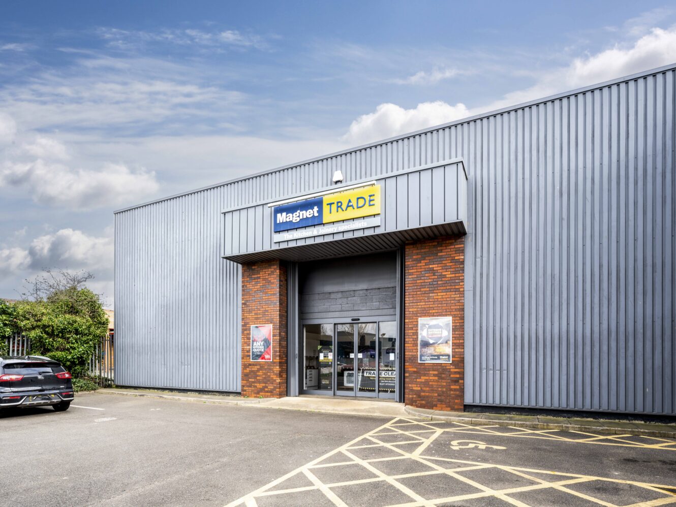 Exterior view of a Magnet Trade store with a metal facade, glass entrance doors, brick accents, and cars parked outside in the parking area.