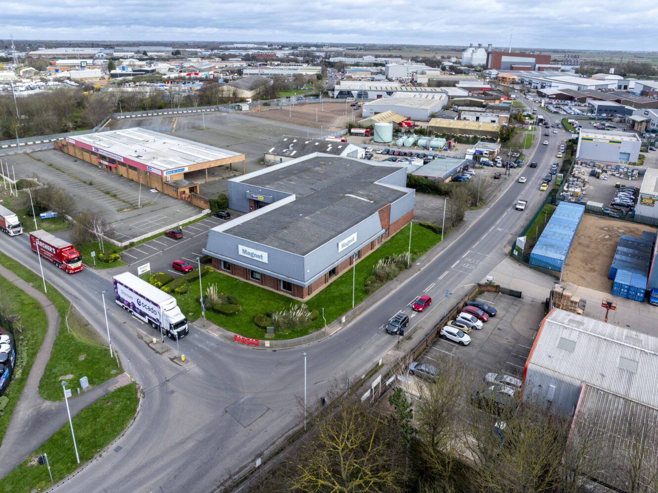 Aerial view of an industrial area with warehouses, trucks on the road, parked cars, and adjacent vacant parking lots under a cloudy sky.