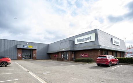 A Magnet store building with a gray exterior, large signs, a sale poster in the window, and red cars parked in the lot on a cloudy day.