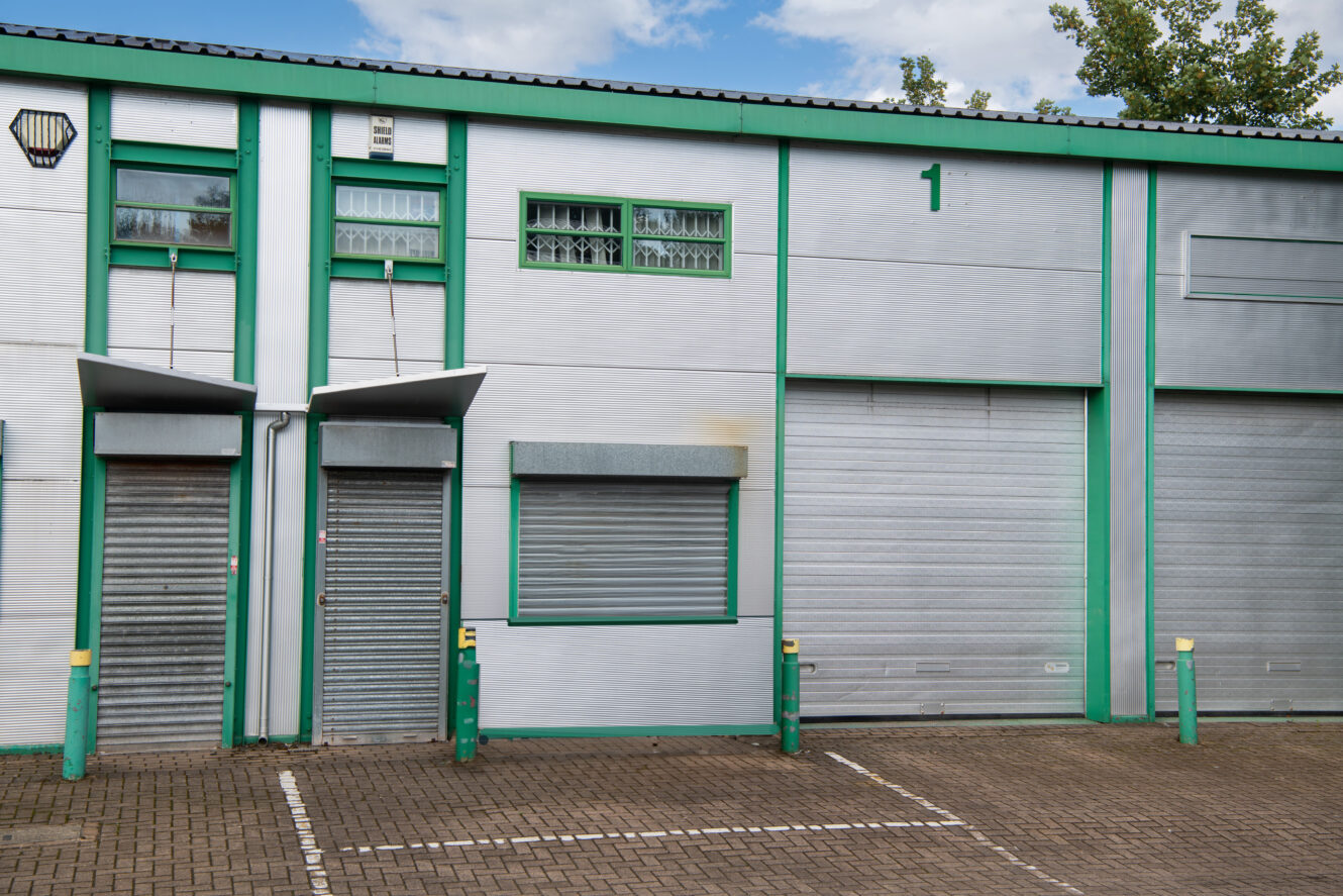 Light gray industrial building with green trim, three metal roller doors (two small, one large), and a numbered 1 above the large door; empty parking spaces in front.
