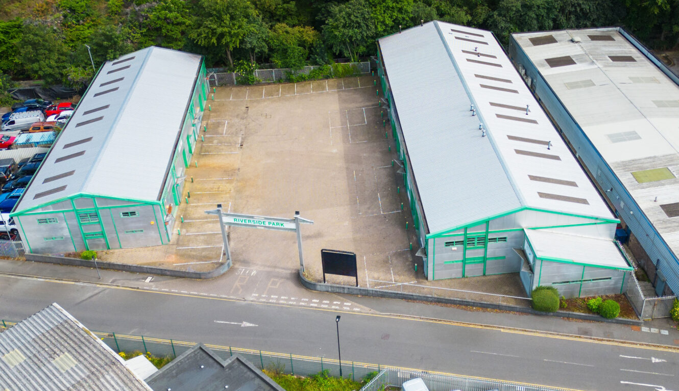 Aerial view of two industrial warehouse buildings with green trim facing each other, separated by a fenced courtyard labeled Riverside Park, next to a street and trees.