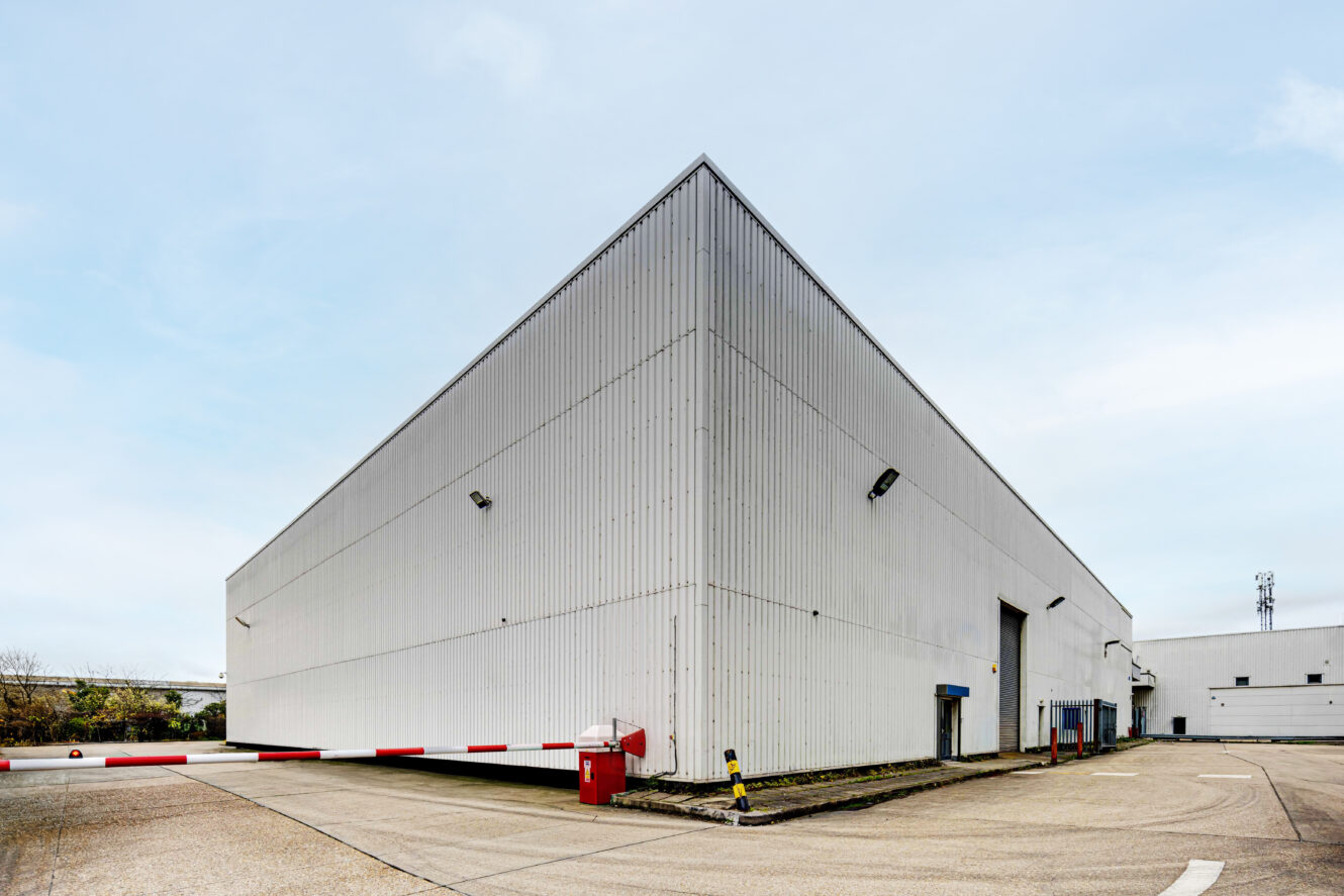 Large industrial warehouse with white metal siding, surrounded by a paved lot, red and white barriers, and minimal landscaping under a cloudy sky.