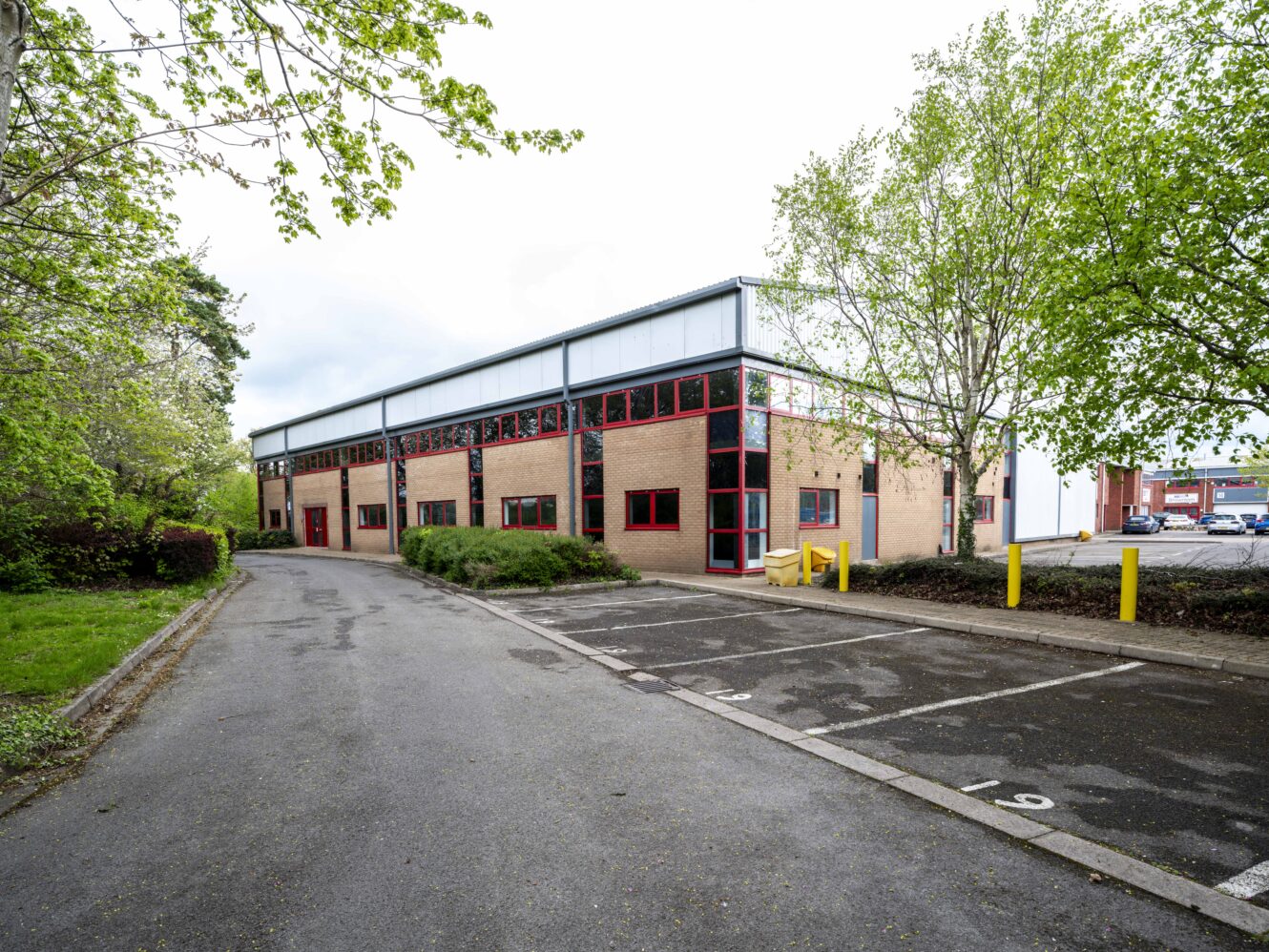 A modern brick office building with red window frames, adjacent to a small parking lot with numbered spaces, surrounded by trees.