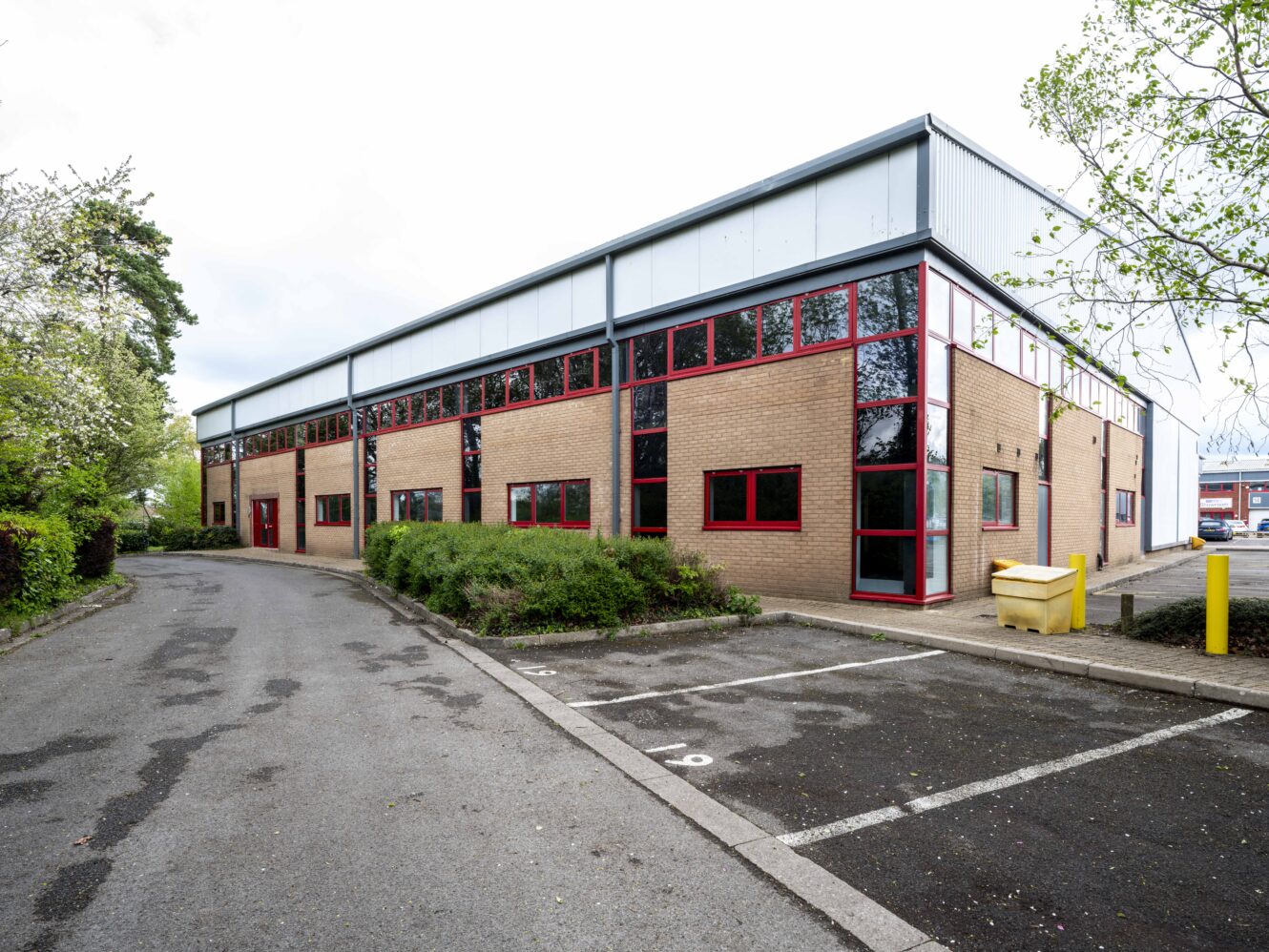 A modern commercial or industrial brick building with red-framed windows, surrounded by greenery, parking spaces, and a paved driveway.