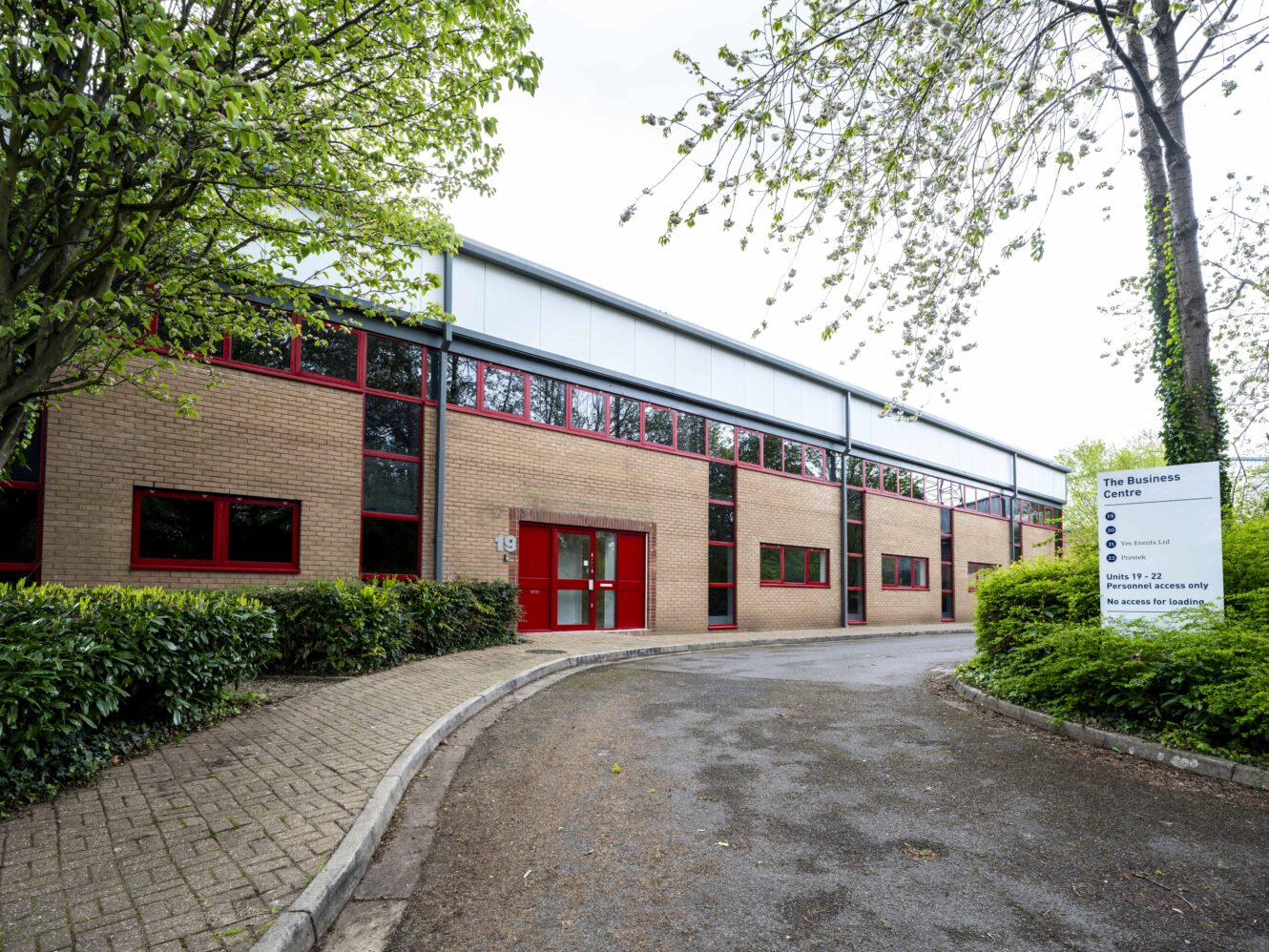 A modern, two-story office building with red-trimmed windows, surrounded by bushes and trees. A sign labeled The Business Centre stands near the entrance.
