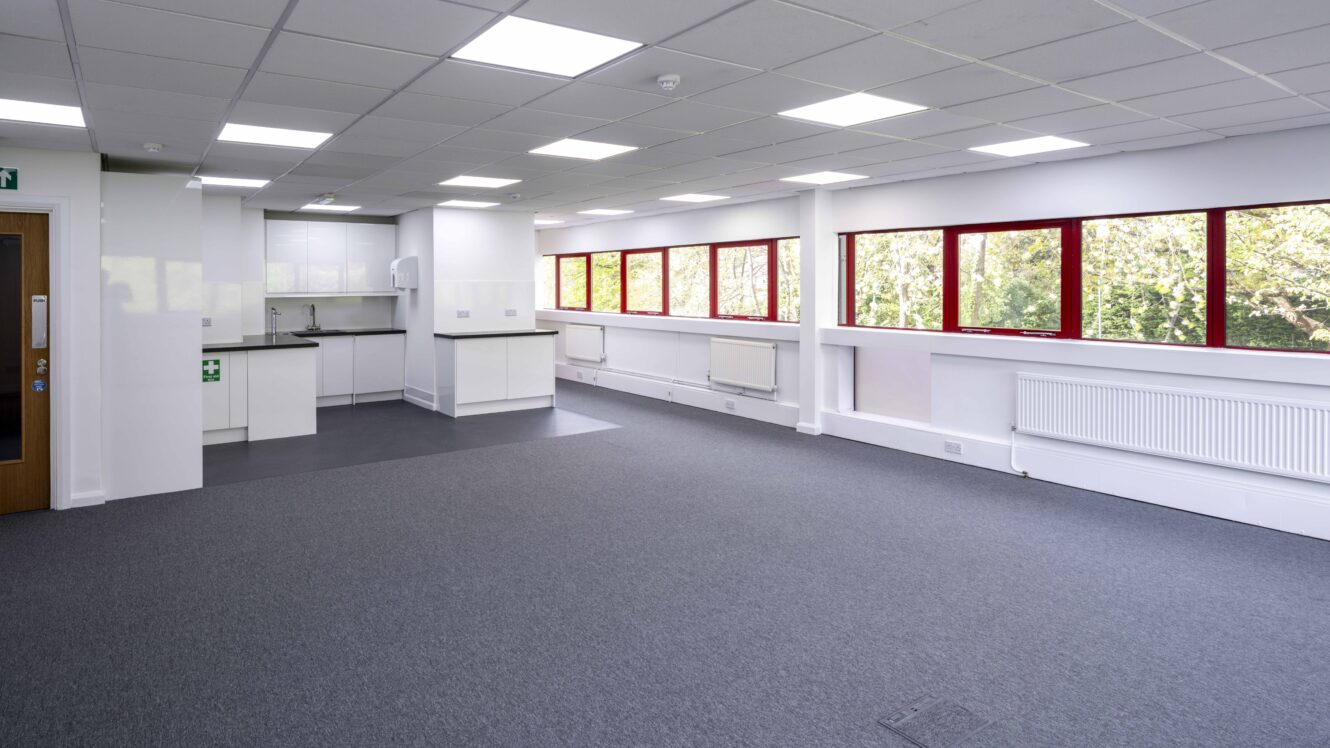 A bright, empty office room with white walls, grey carpet, red-trimmed windows, and a small kitchenette in one corner.