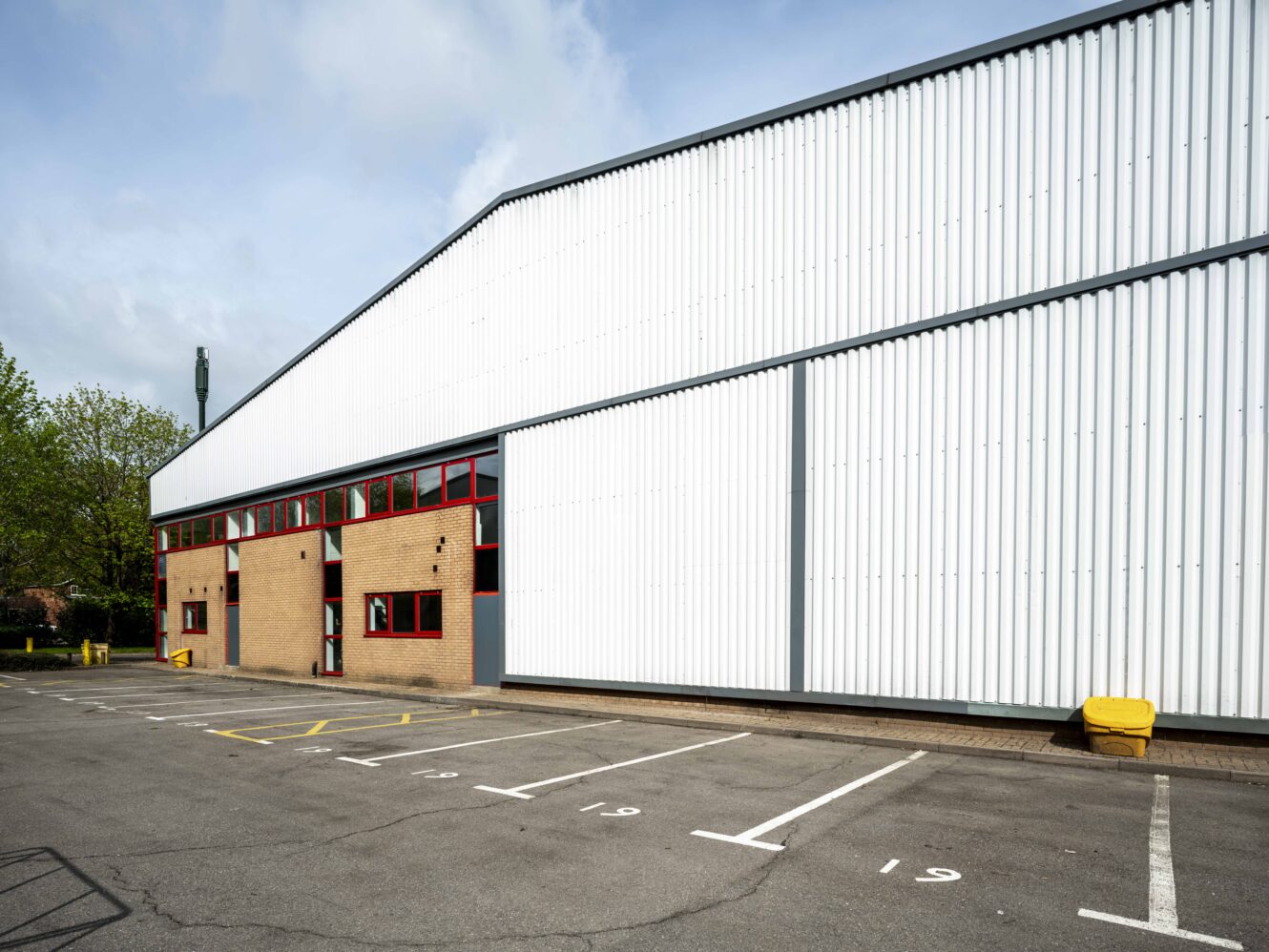 Large industrial warehouse with white corrugated metal siding, brick lower facade, red-framed windows, and an empty parking lot in front.