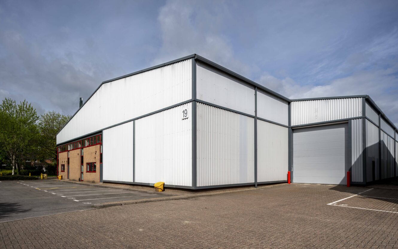 A large, white industrial warehouse with a roller shutter door and numbered sign, surrounded by a paved parking lot and some greenery in the background.