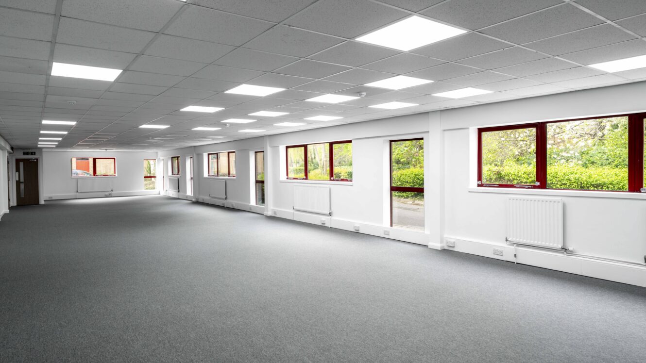Spacious empty office with gray carpet, white walls, ceiling lights, and large windows overlooking greenery.