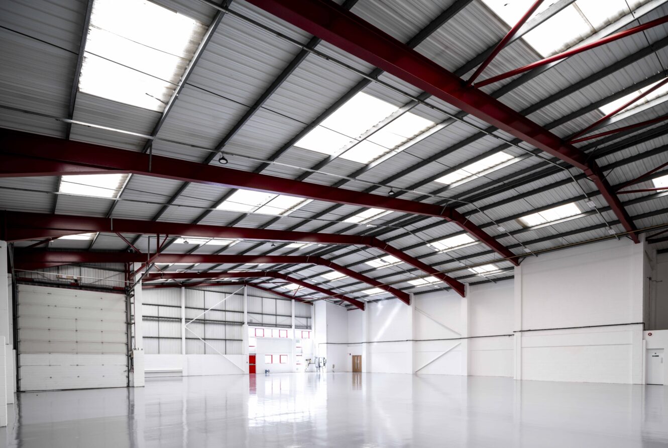 Interior of a large, empty industrial warehouse with white walls, a shiny floor, red steel beams, and skylights in the metal ceiling.