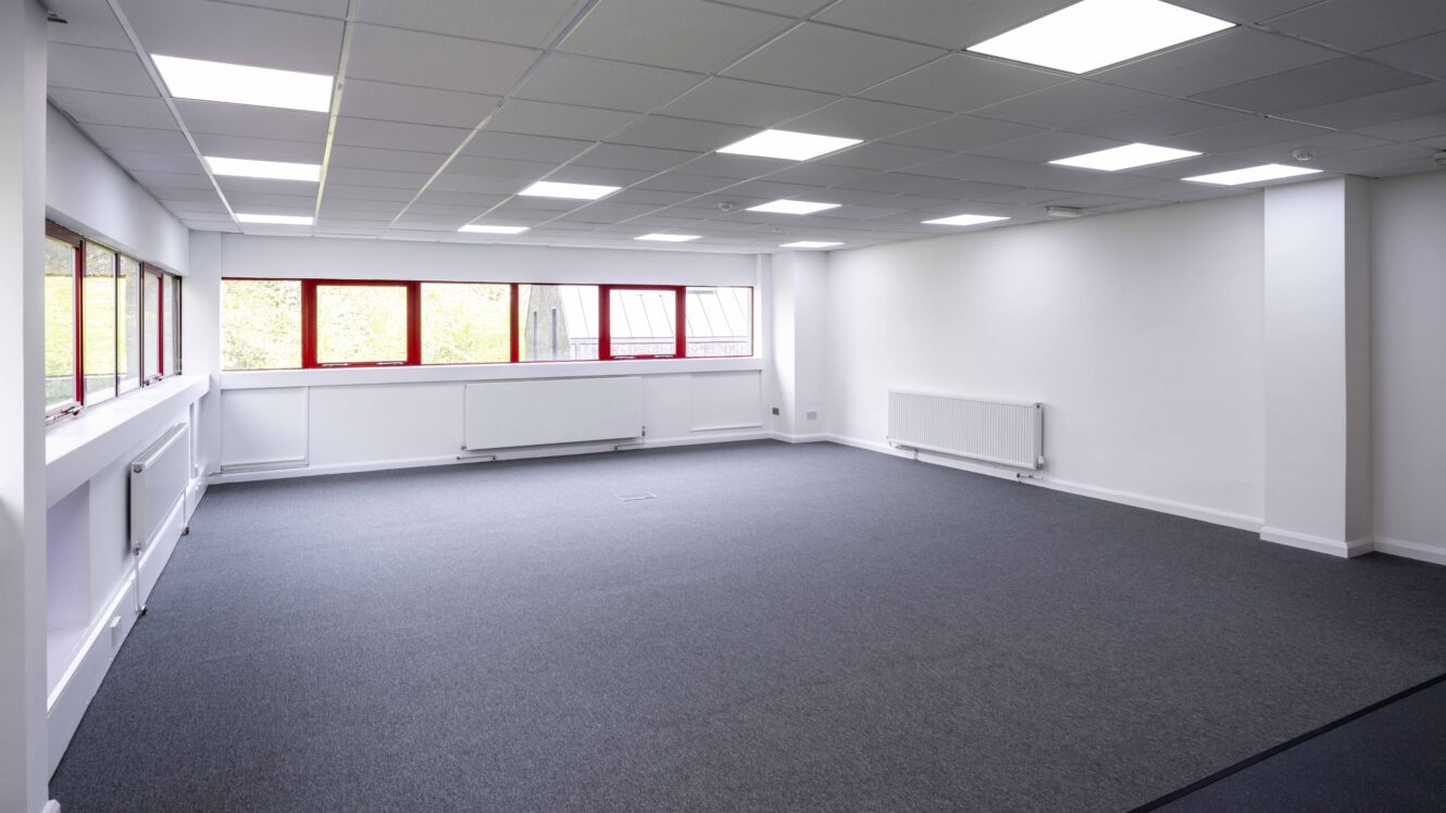An empty office space with gray carpet, white walls, ceiling lights, large windows, and wall-mounted radiators.