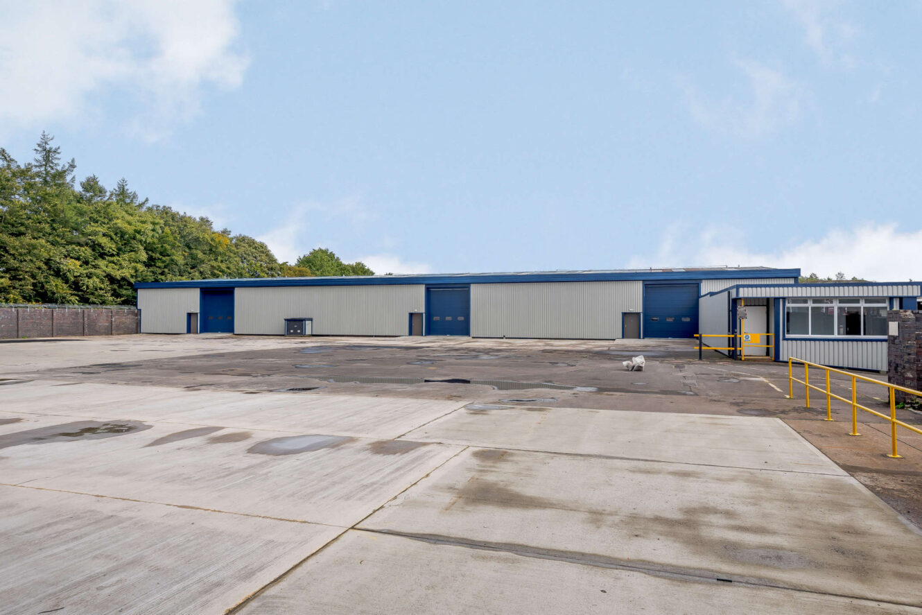 Large industrial warehouse with blue doors and a wide empty concrete lot in front, bordered by trees and a security fence on the left.