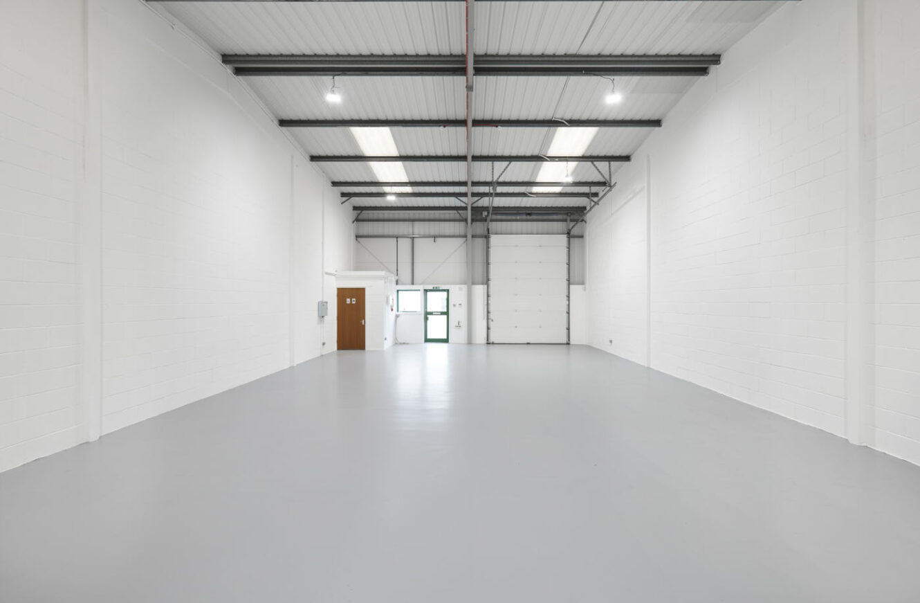 An empty industrial warehouse unit with white walls, a high ceiling, exposed beams, and a gray concrete floor. Fluorescent lights hang from the ceiling. A door and window are visible at the far end.