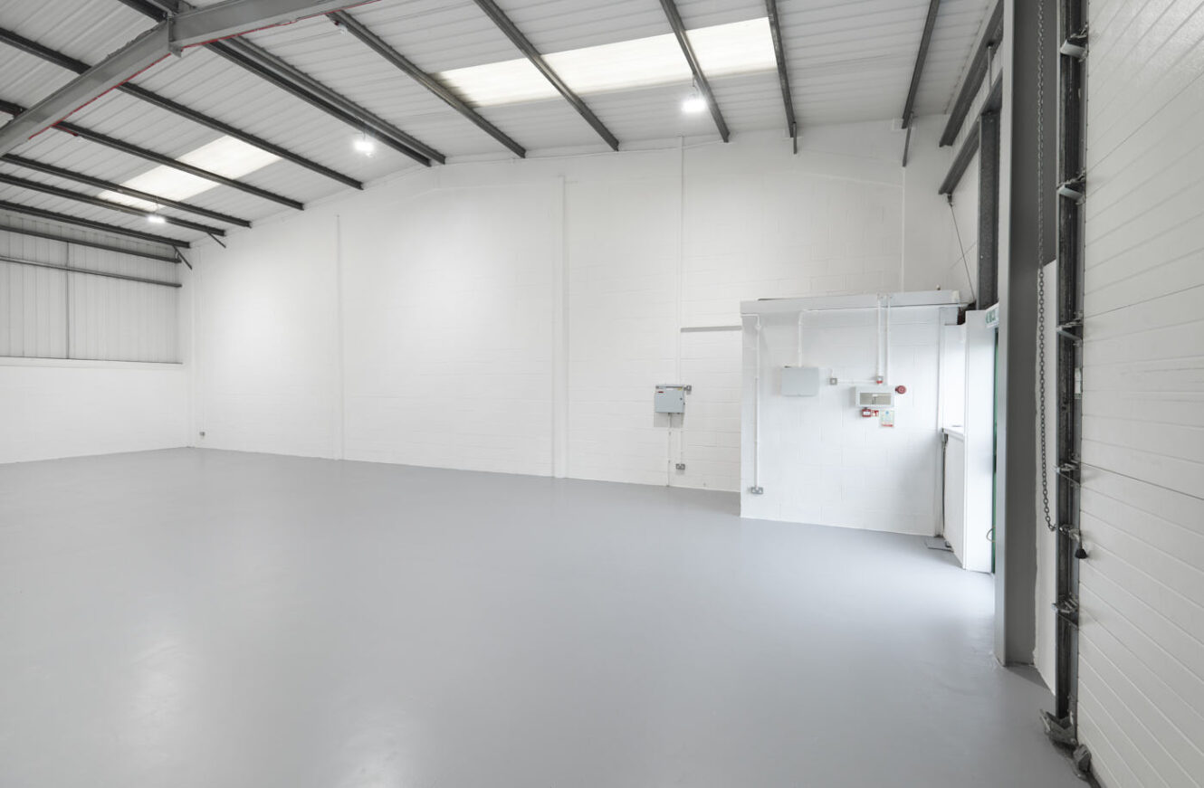 Empty industrial warehouse with white walls, high ceilings, exposed beams, and gray flooring. Fluorescent lights and electrical panels are visible on the back wall near a door.