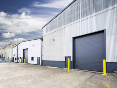Exterior view of a modern industrial warehouse with metal siding, two large roller doors, and a paved loading area under a partly cloudy sky.