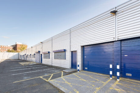 Row of industrial storage units with blue doors and white siding, adjacent to an empty parking lot with marked spaces and yellow-striped accessible parking areas.