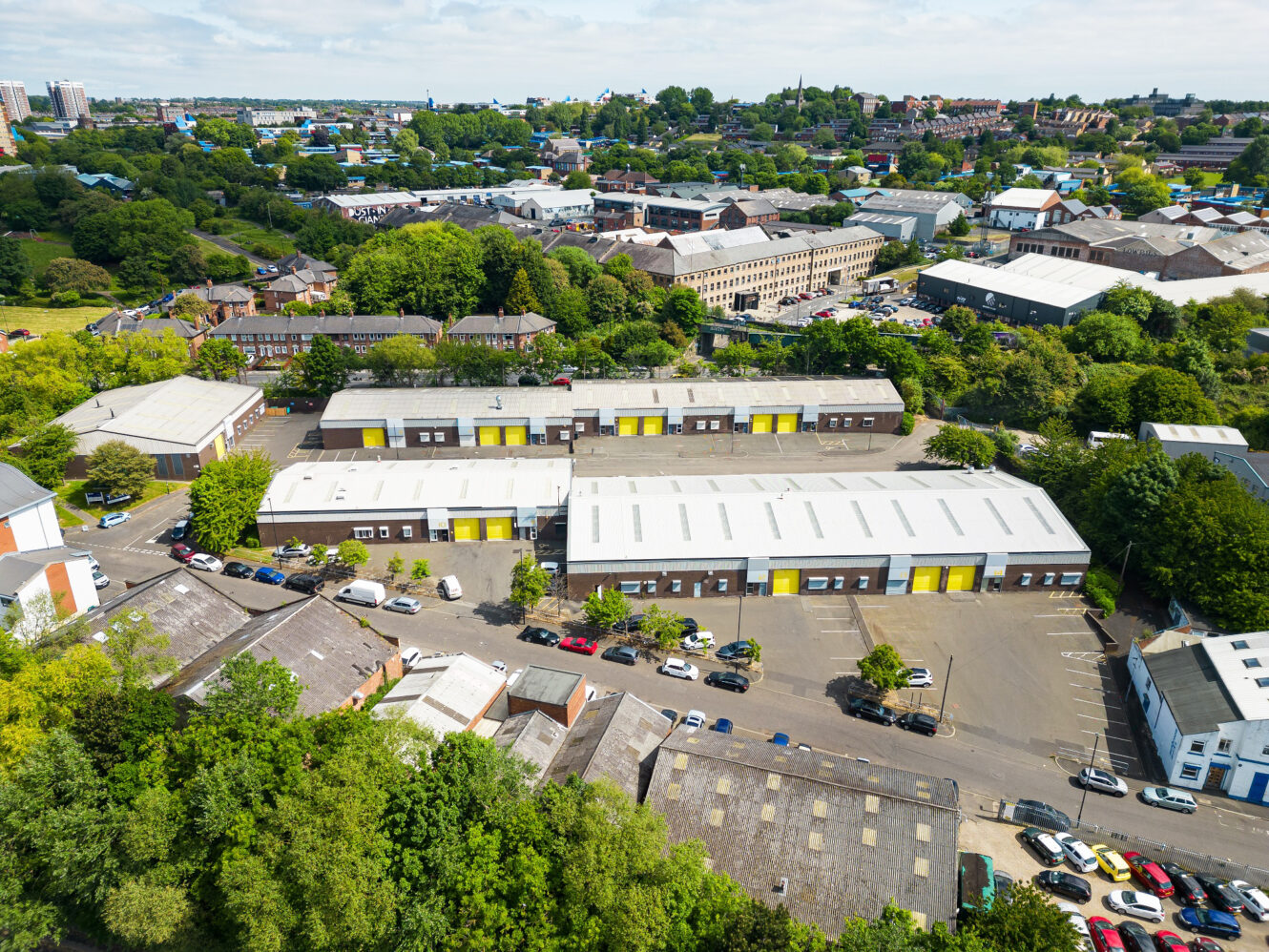 Aerial view of an industrial estate with multiple warehouse buildings featuring yellow doors, surrounded by trees and neighboring city structures.