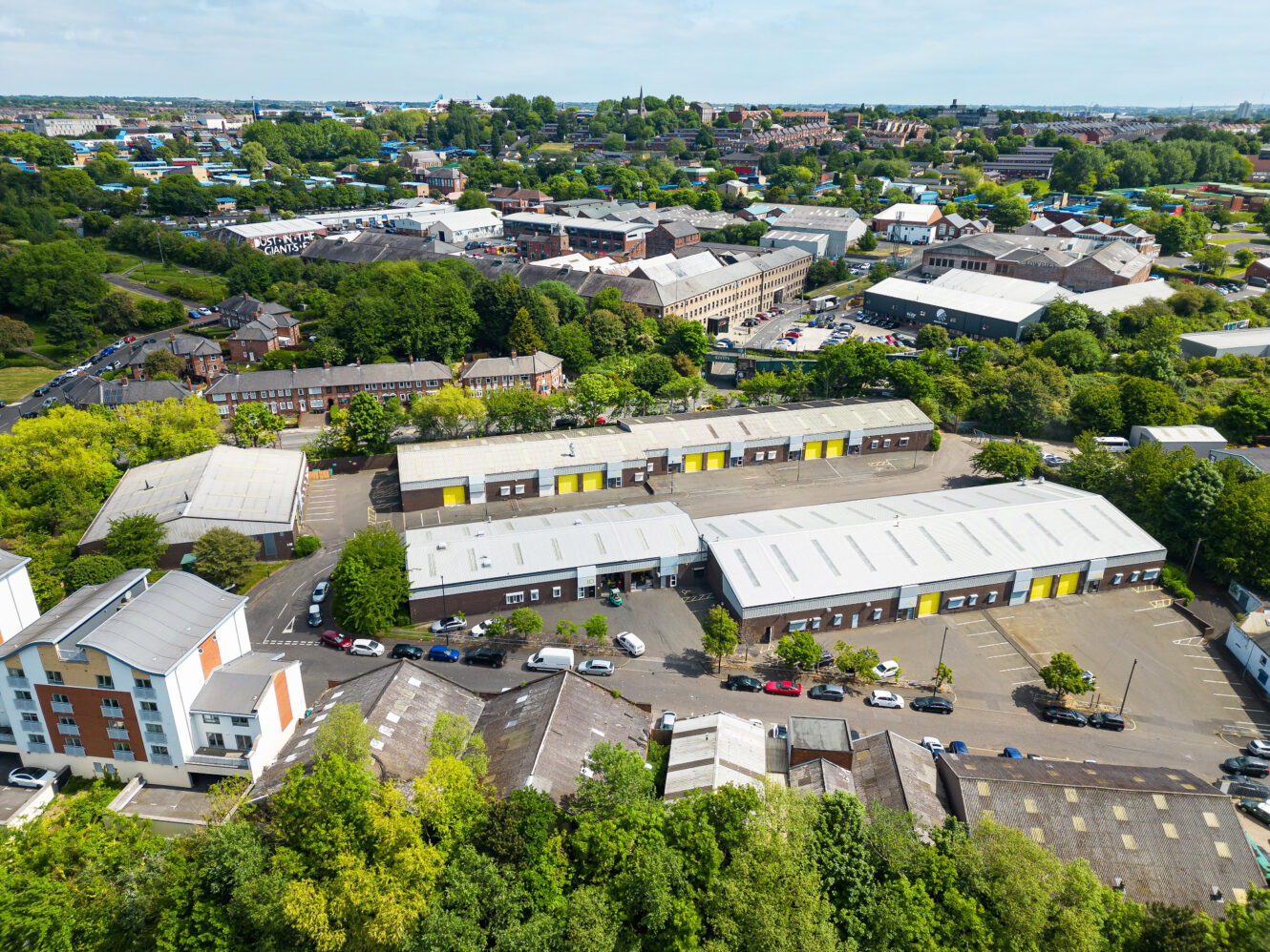 Aerial view of an industrial estate with several warehouses, yellow doors, parked cars, surrounding trees, and residential buildings in the background.