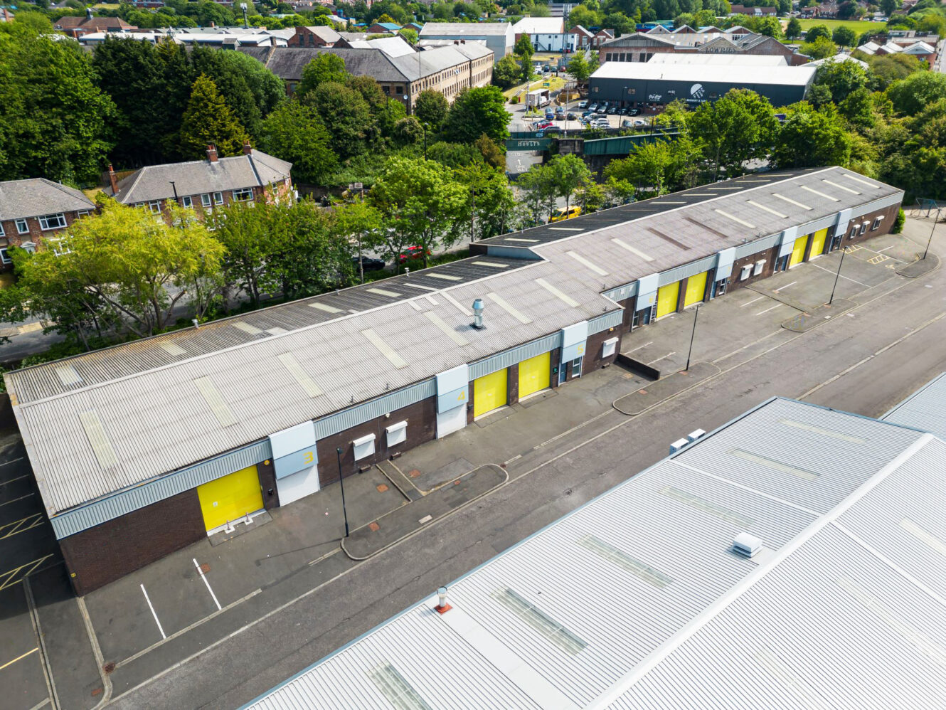 Aerial view of an industrial building with multiple yellow roller doors and adjacent parking spaces, surrounded by trees and other buildings.