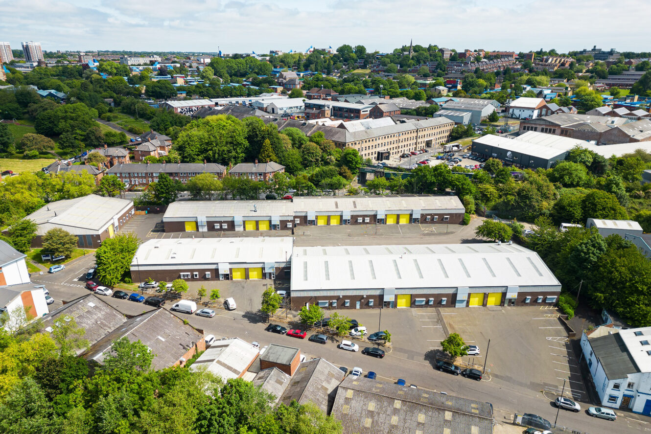 Aerial view of an industrial estate with several warehouse buildings featuring yellow doors, surrounded by parked cars, trees, and a residential area in the background.