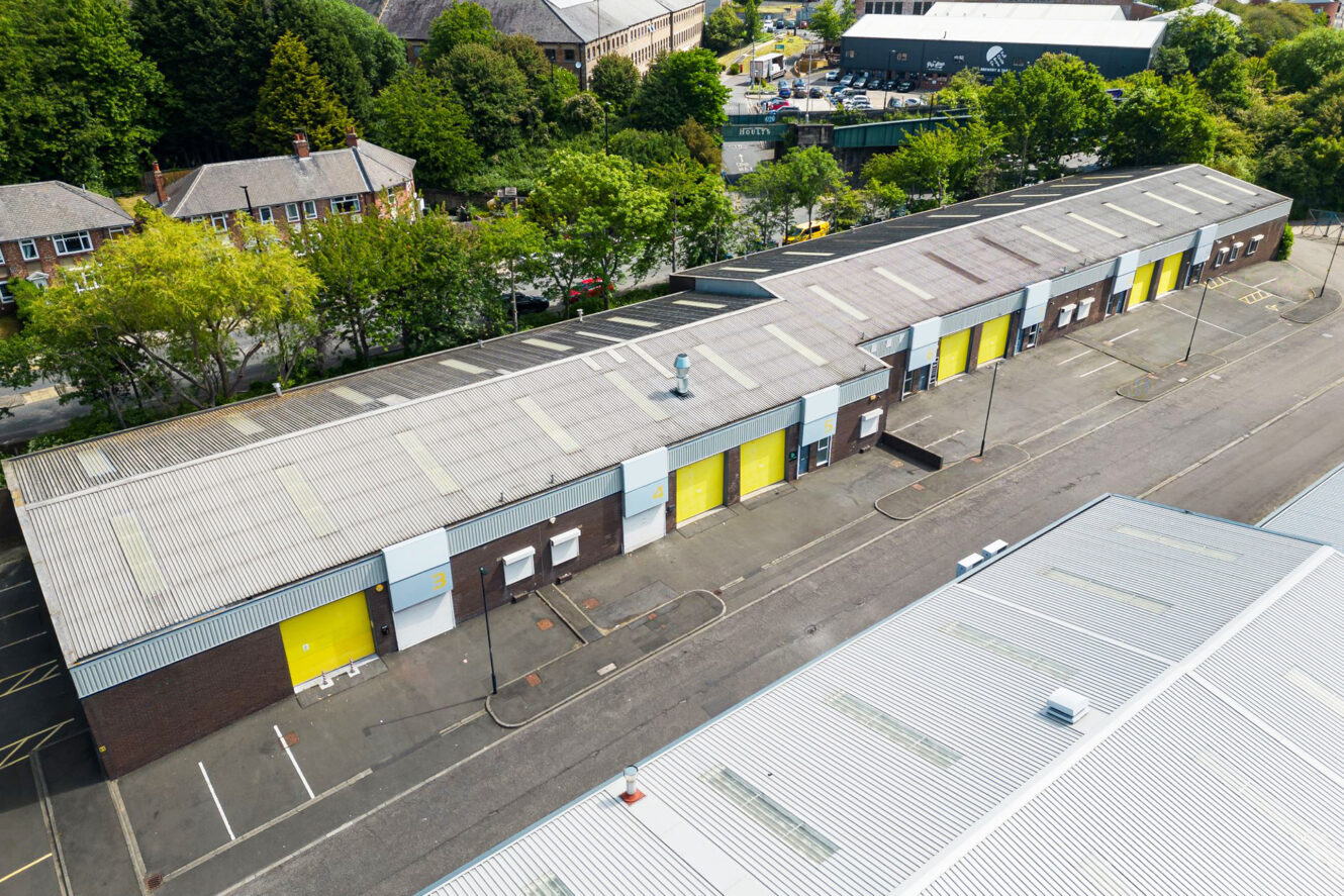 Aerial view of a row of industrial warehouse units with yellow doors and a surrounding asphalt parking area, bordered by trees and adjacent buildings.