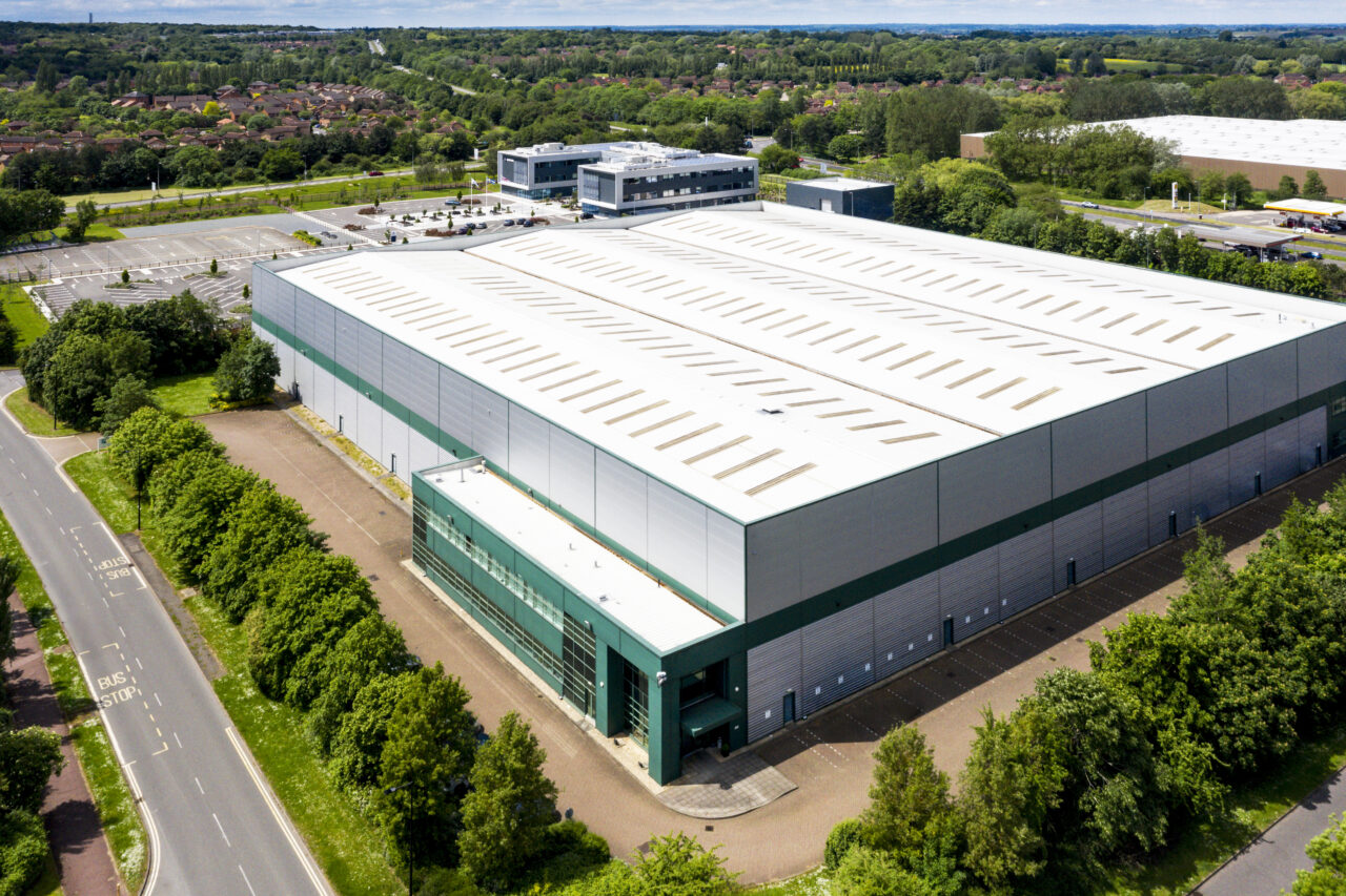 Aerial view of a large industrial warehouse with a white roof, surrounded by trees, parking lots, and adjacent roads in a suburban area.
