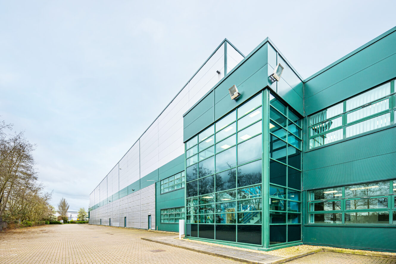 Modern industrial warehouse building with large glass entrance, green and gray exterior panels, and an empty paved area in the foreground.