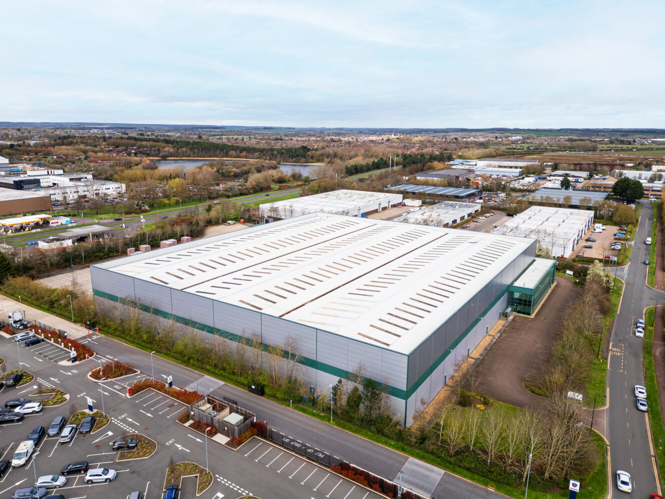 Aerial view of a large industrial warehouse building with a white roof, surrounded by parking lots, roads, and other commercial structures.