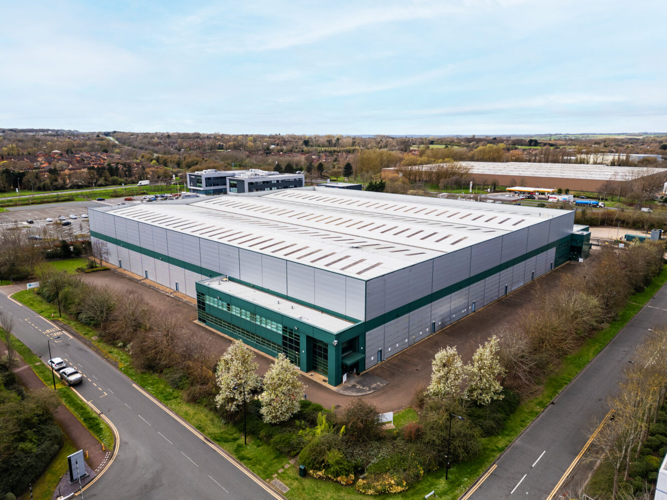 Aerial view of a large, modern industrial warehouse with gray and green exterior, surrounded by roads, trees, and parking areas.
