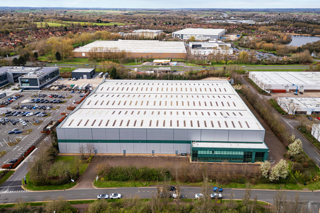 Aerial view of a large warehouse building with adjacent parking lot, surrounded by roads, trees, and other industrial buildings in a semi-rural area.
