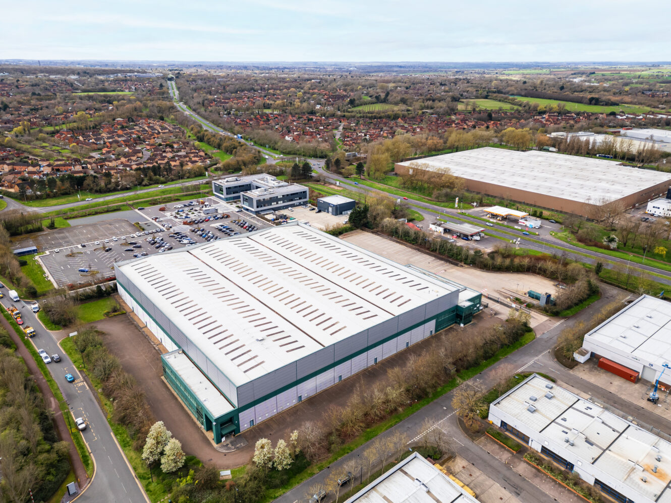Aerial view of a large industrial warehouse with adjacent parking lots, surrounded by trees, roads, and other buildings in a suburban area.