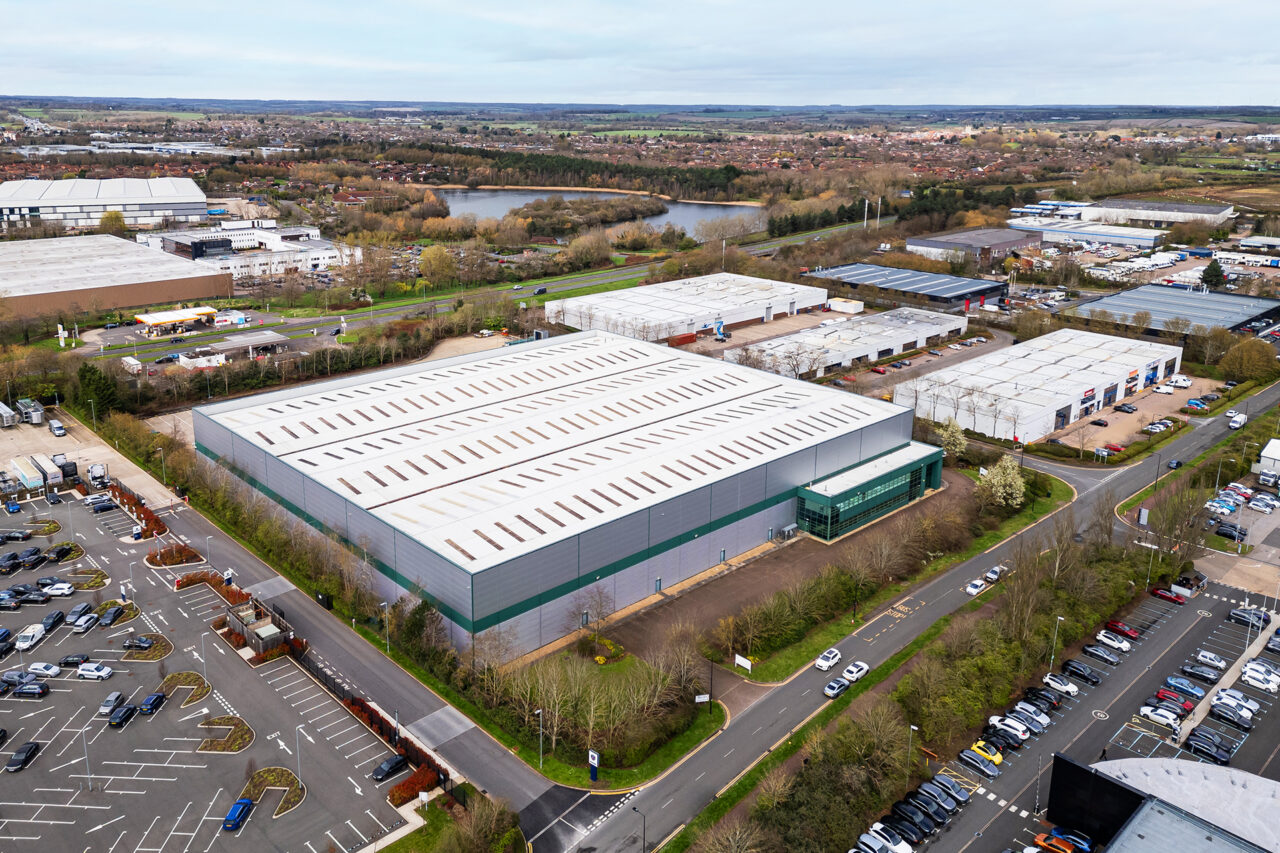 Aerial view of a large industrial warehouse with a white roof, surrounded by parking lots, trees, and other commercial buildings in an industrial area.