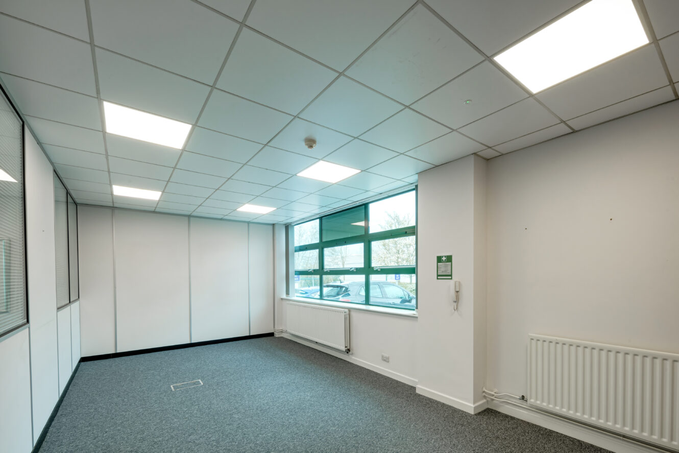 Empty office room with gray carpet, white walls, large window, ceiling lights, and a radiator under the window.