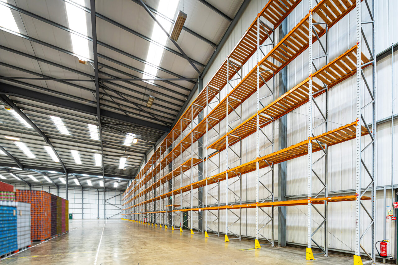 Empty industrial warehouse with tall, empty metal storage racks along one wall and stacks of goods in the background. The space is brightly lit with a high ceiling.