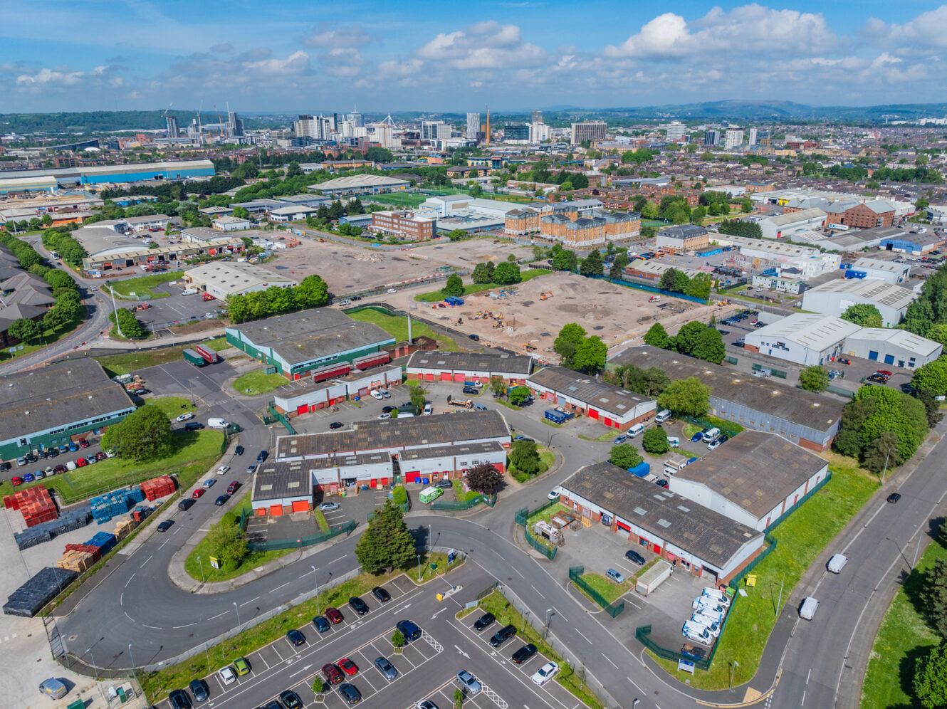 Aerial view of an industrial estate with warehouses, parking lots, and roads, set against a cityscape background under a partly cloudy sky.