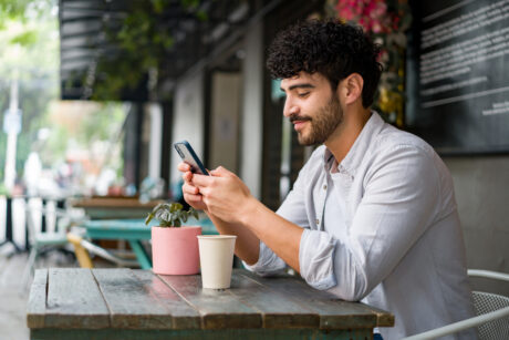 A man sits at an outdoor café table, looking at his phone. A plant in a pink pot and a disposable coffee cup are on the table.