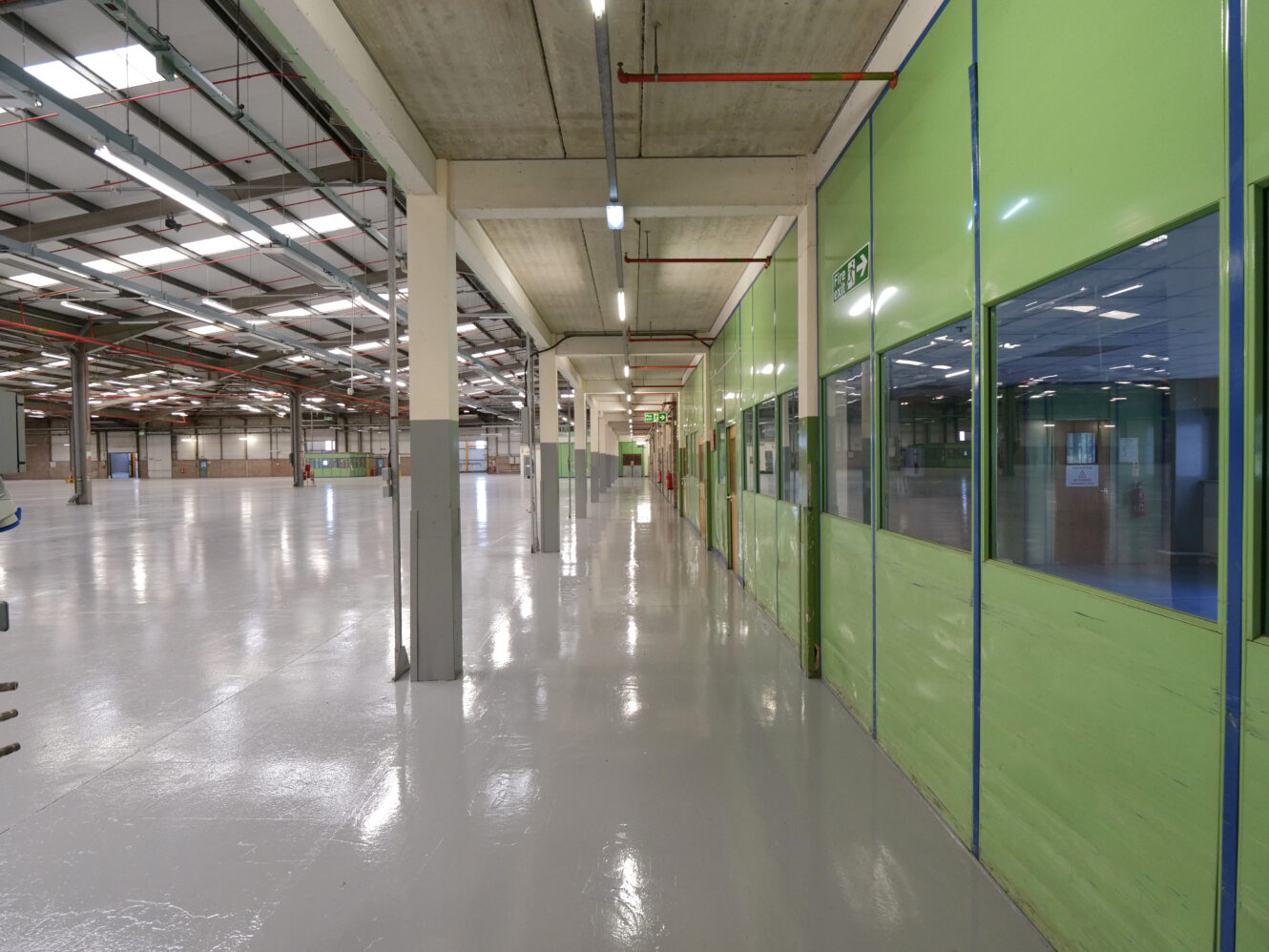 Wide view of an empty industrial warehouse interior with green partition walls, large windows, polished floors, and overhead lighting.