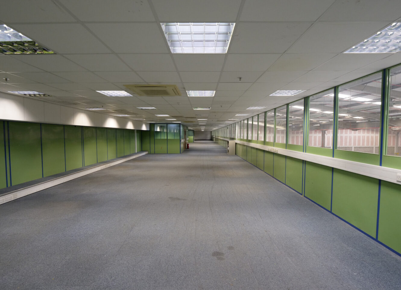 A long, empty office corridor with green partition walls, carpeted floor, and fluorescent ceiling lights, stretching into the distance.