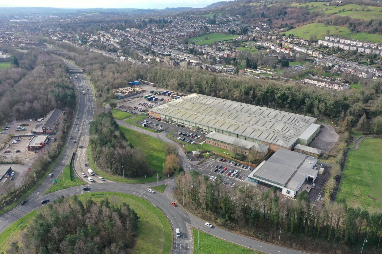 Aerial view of a large industrial building surrounded by trees, parking lots, and roads, with a residential neighborhood and hills in the background.