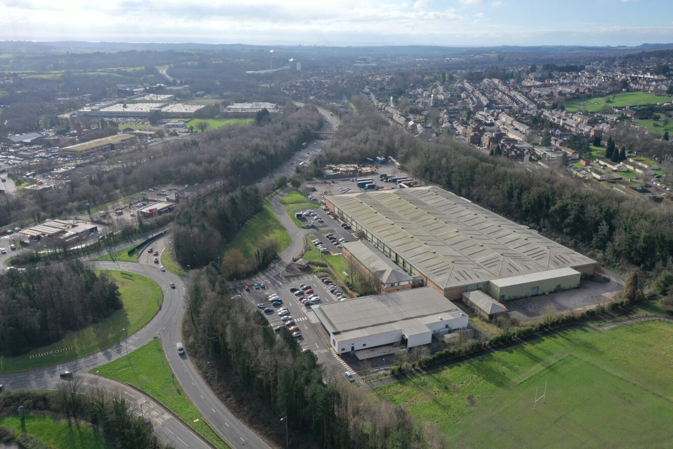 Aerial view of a large industrial warehouse complex surrounded by trees, parking lots, and roads, with a residential area visible in the background.