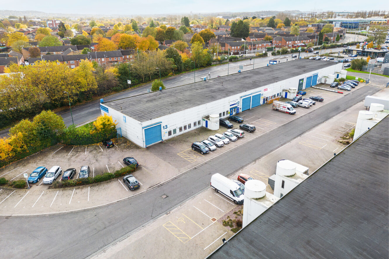 Aerial view of a warehouse with multiple blue doors, several parked cars, and vans in the parking lot, surrounded by trees and nearby residential houses.