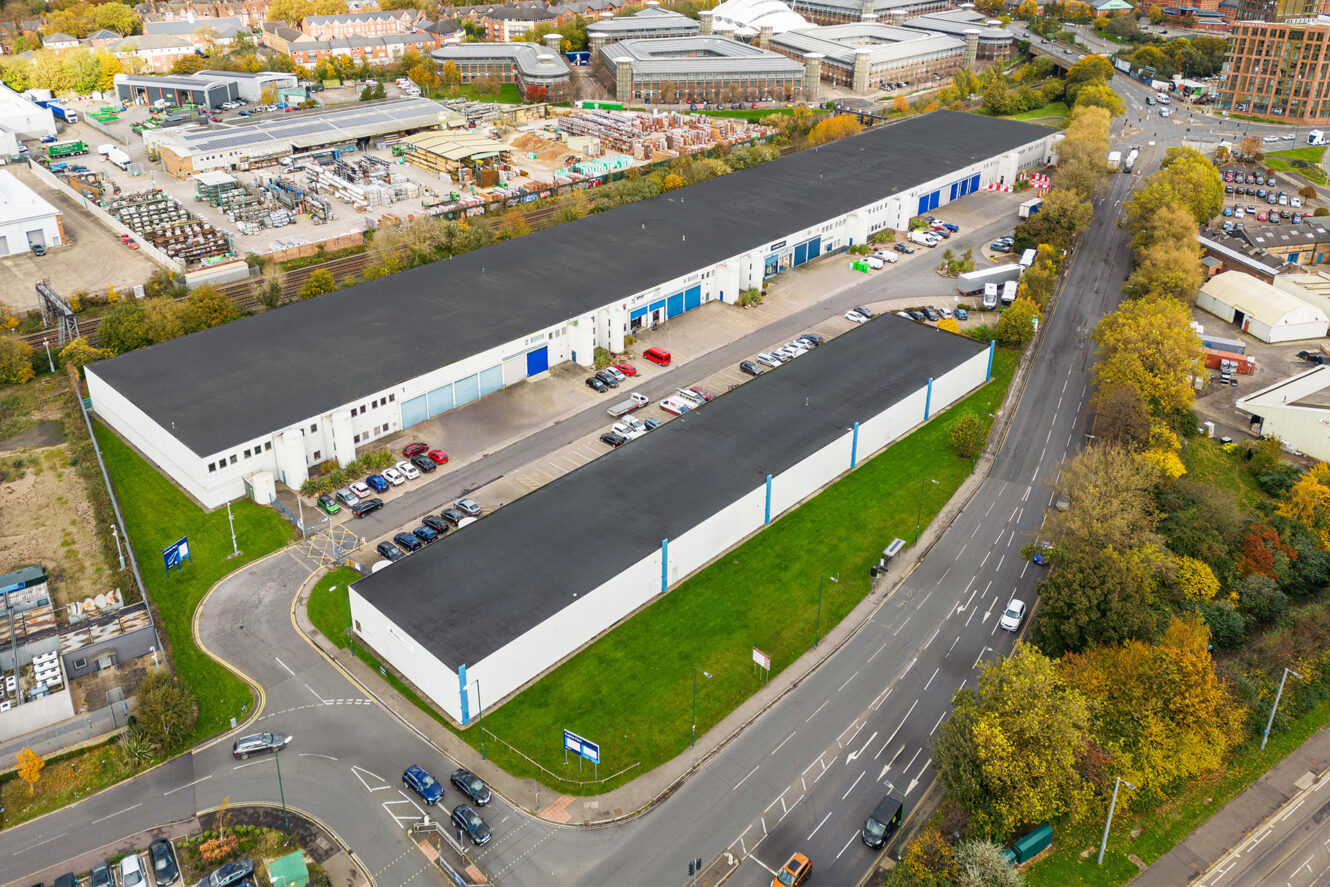 Aerial view of an industrial complex with two large warehouse buildings, surrounding parking lots with cars, and adjacent roads bordered by green lawns and trees.