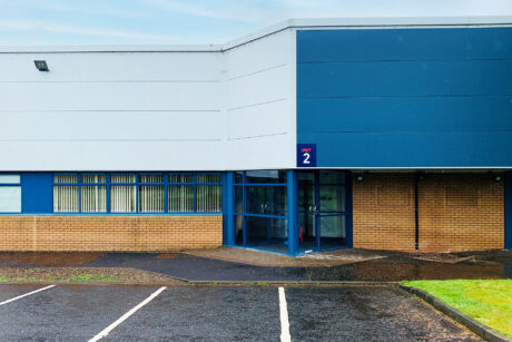 A modern building with blue and white panels, brick lower walls, large windows, and an entrance marked Unit 2 next to an empty parking lot.