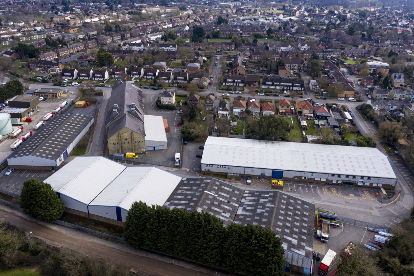 Aerial view of an industrial area with warehouses in the foreground and residential houses in the background, separated by a row of trees.