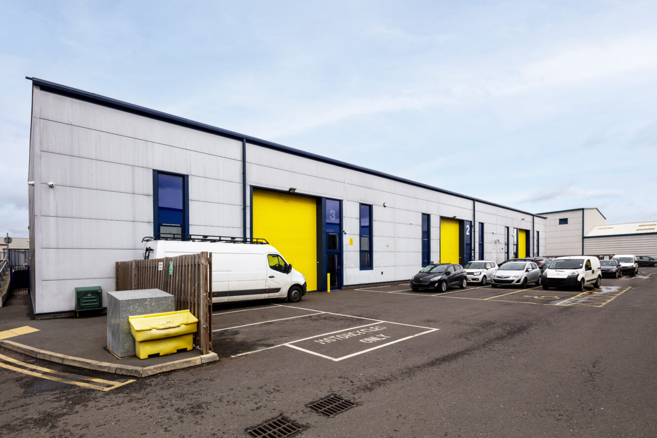 A row of industrial warehouse units with yellow accent doors, parked cars, a white van, and a yellow dumpster in the parking lot under a cloudy sky.