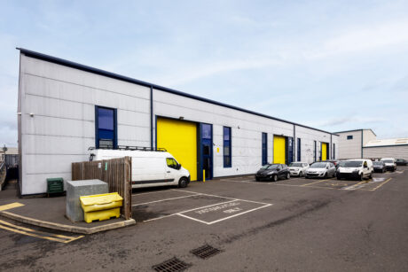 A row of industrial warehouse units with yellow accent doors, parked cars, a white van, and a yellow dumpster in the parking lot under a cloudy sky.
