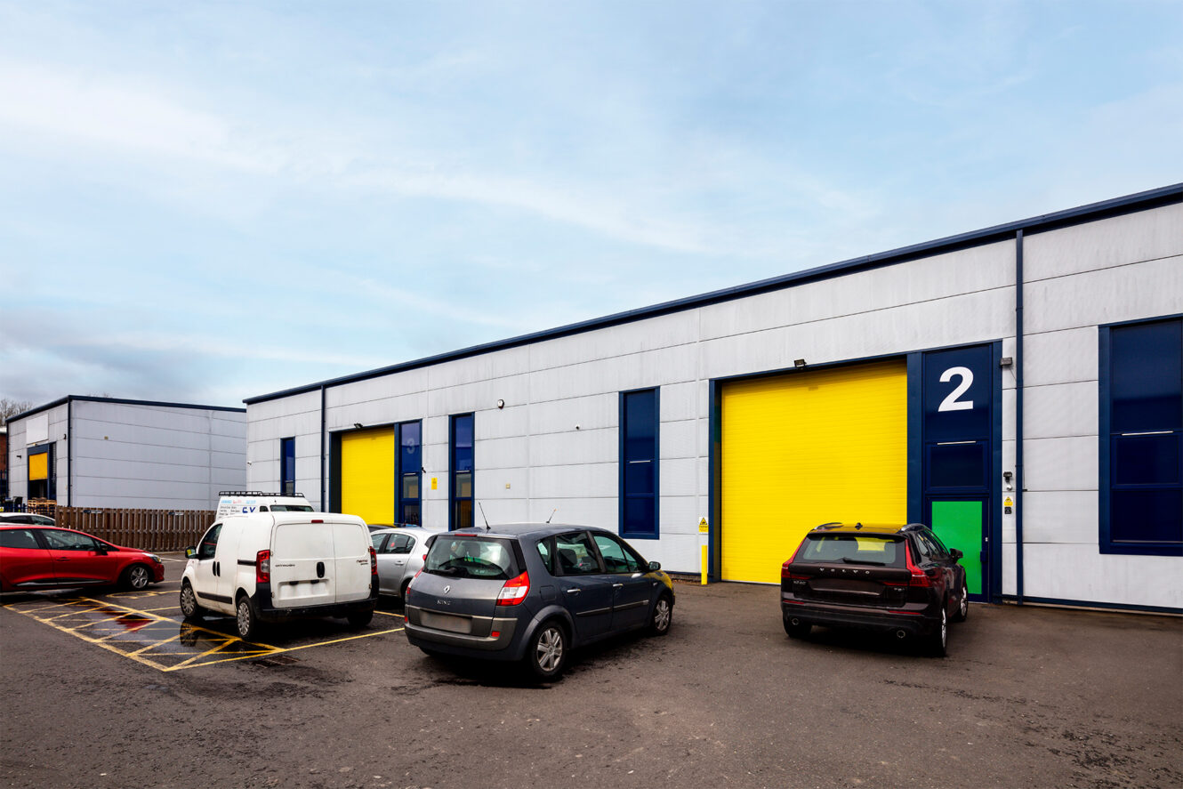 Several cars are parked outside a row of industrial warehouse units with bright yellow shutters and blue accents. One unit is labeled with a large number 2.
